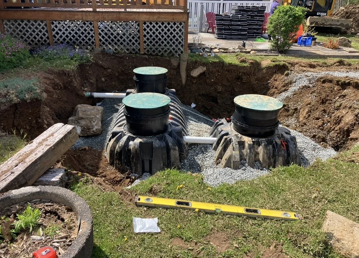 Underground septic tank system installation in a backyard with gravel and large black plastic tanks connected by white pipes, surrounded by grass and construction tools.
