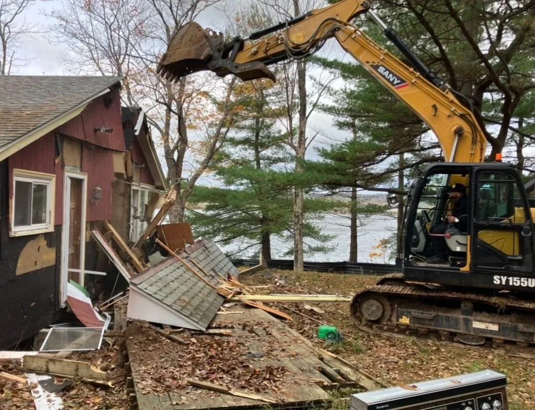 A yellow excavator tearing off the side of a house with trees and a lake in the background.