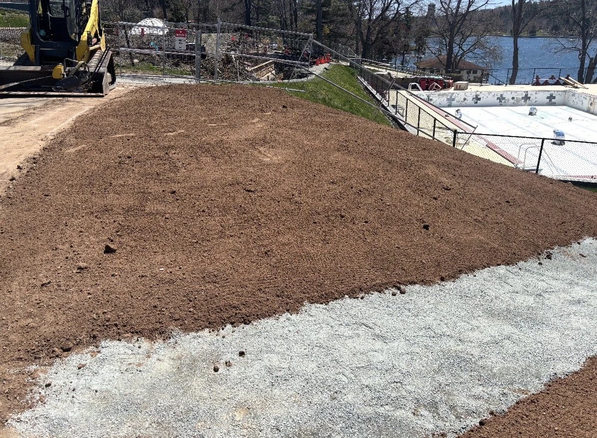 A construction site showing a dirt mound with a dirt path in front, a mini excavator on the left, and a hockey rink and lake in the background.