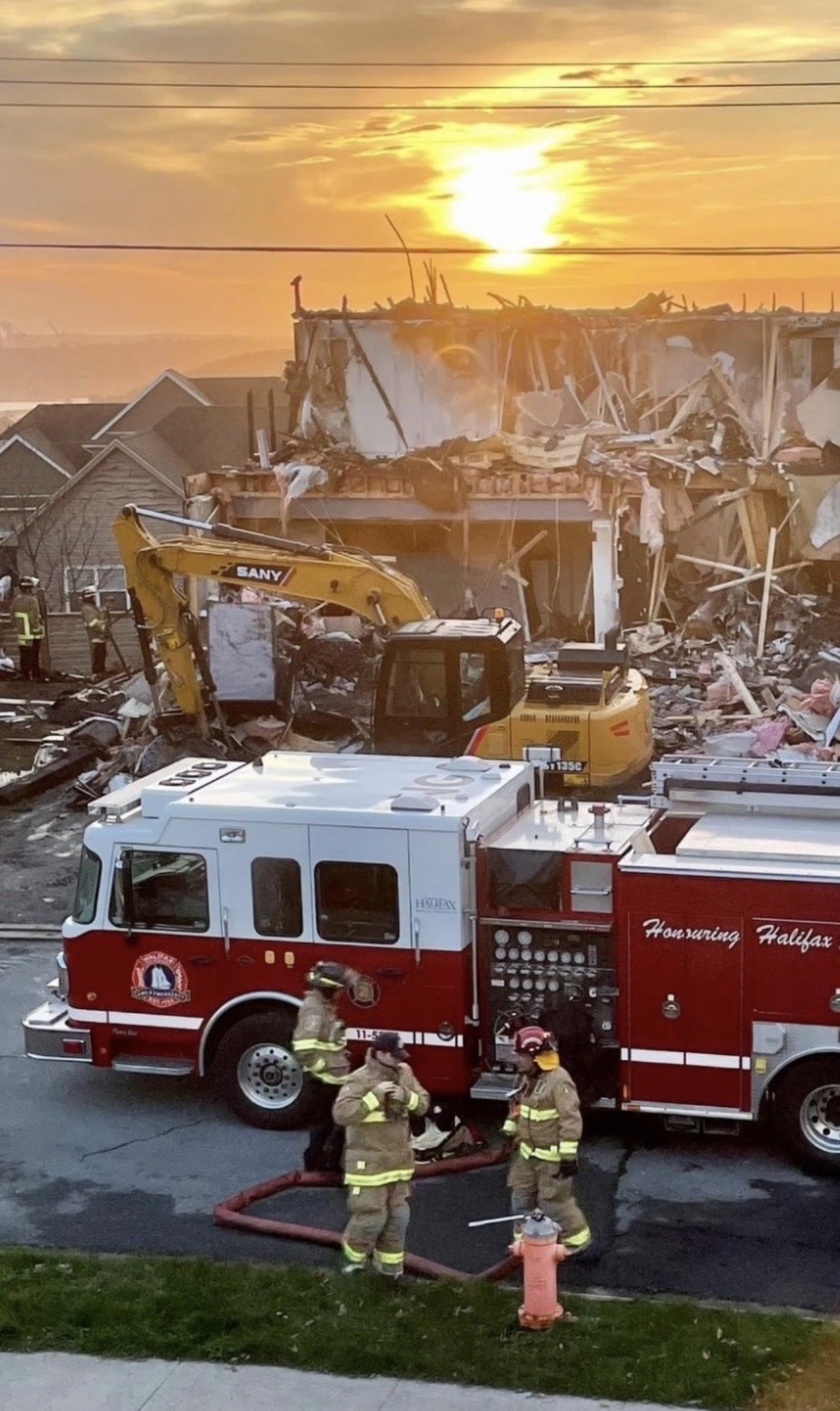 Firefighters work at the scene of a building collapse, with debris and damage visible, during sunset, with a fire truck and rescue equipment present.