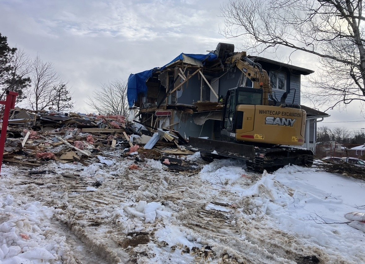 A construction excavator is demolishing a two-story house with debris and snow on the ground.