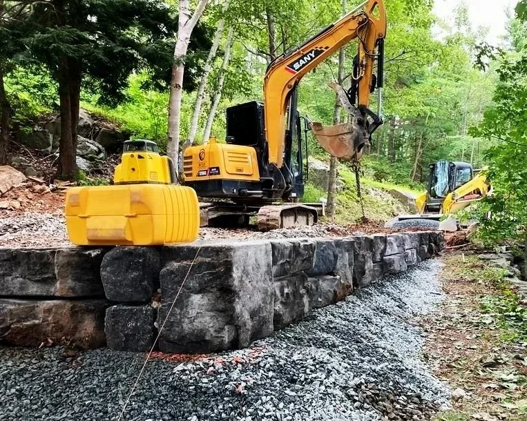 Construction site with two yellow excavators working on a stone wall in a wooded area. One excavator is actively moving dirt, while the other is in the background.