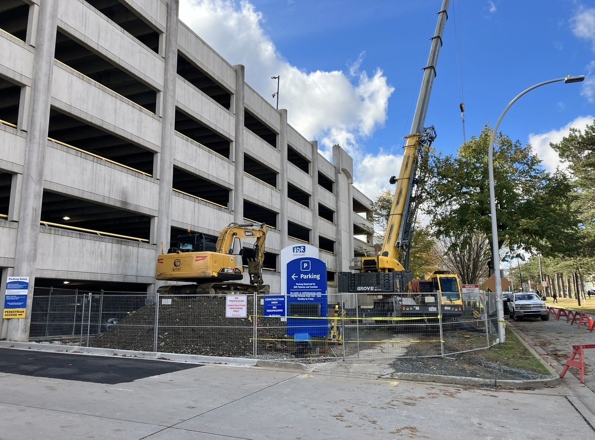 Construction site with a parking garage, a crane, and a small excavator beside a blue parking sign, surrounded by temporary fencing, on a partly cloudy day.