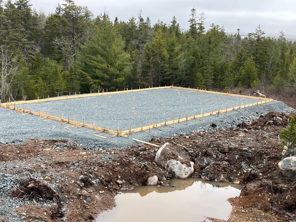 Construction site with gravel and wooden frame, surrounded by trees, with a small pond in the foreground.