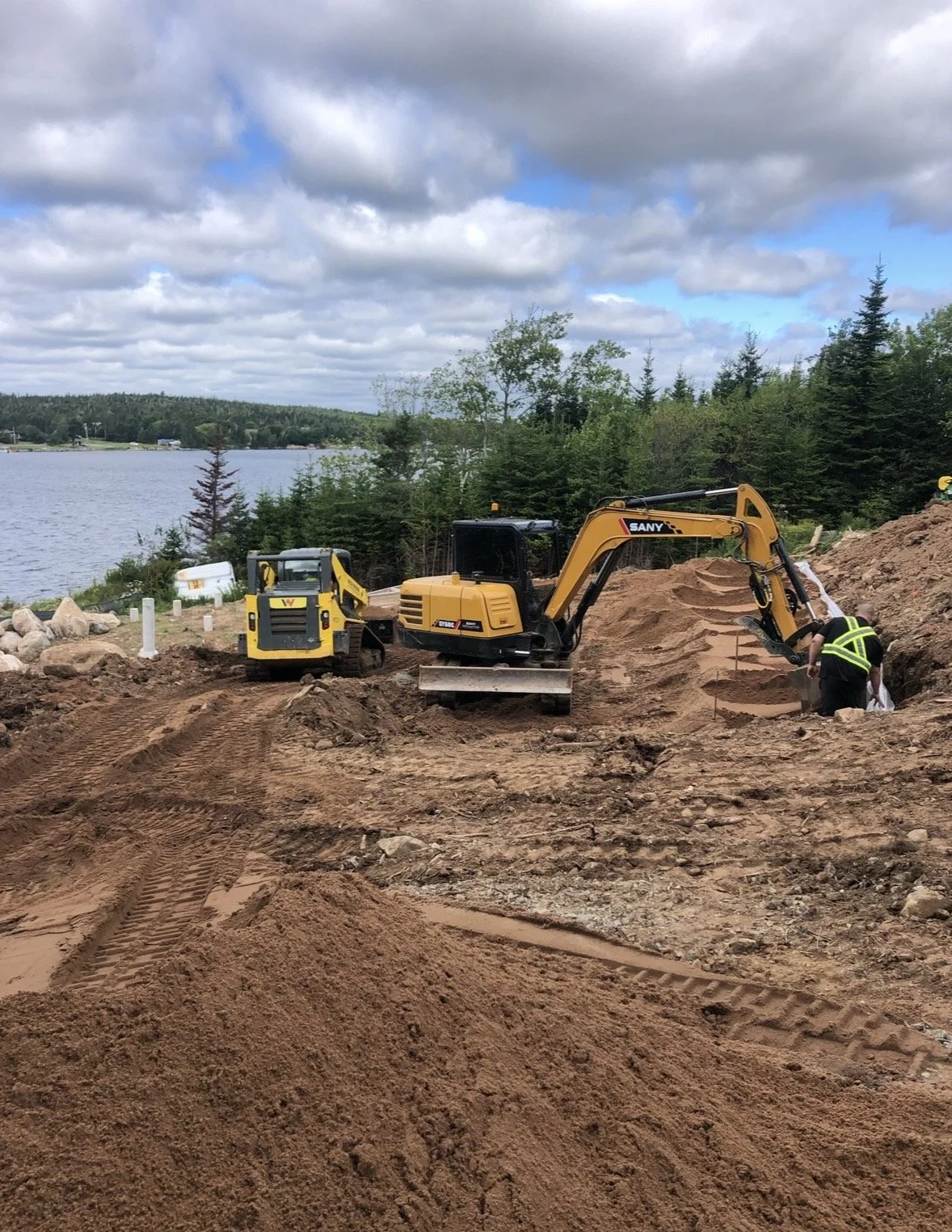 Construction site by a lake with two small yellow excavators, one worker in a high-visibility vest, and a partially dug area with mounds of dirt, trees, and a cloudy sky in the background.
