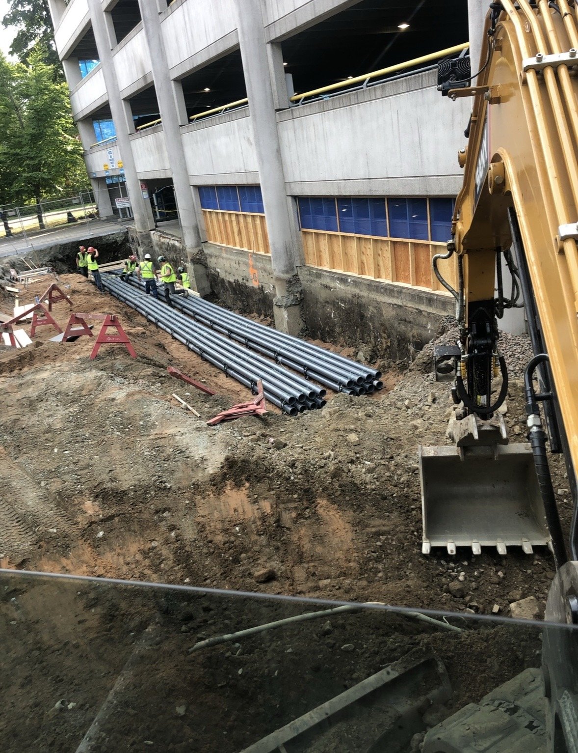 Construction workers installing pipes in a trench outside a multi-story parking garage.