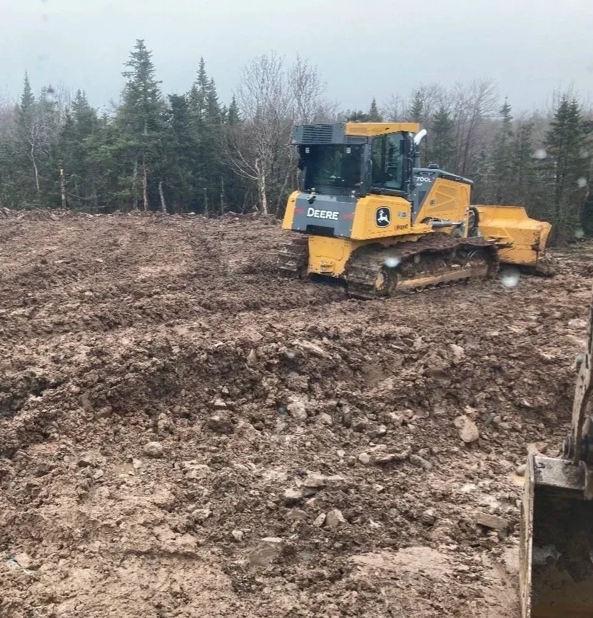 A yellow John Deere bulldozer working on a dirt site with a forest of trees in the background.
