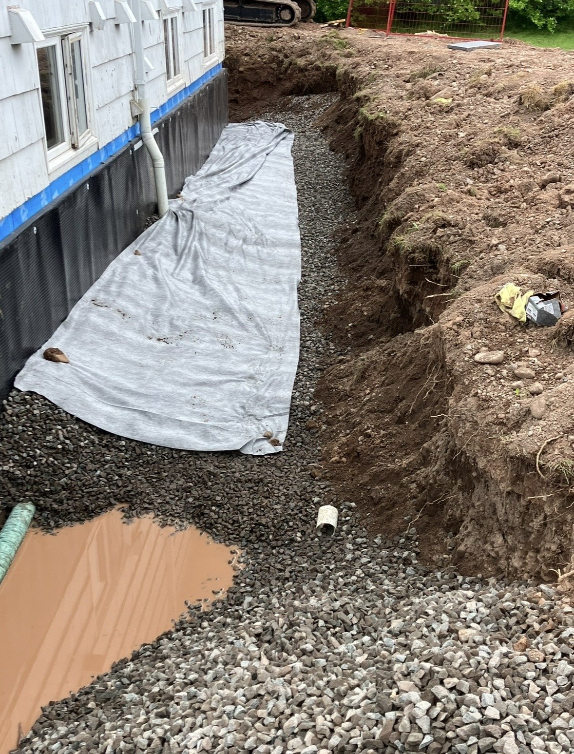 A house undergoing foundation work with a trench dug around the perimeter. A sheet of plastic or tarp is laid down over gravel in the trench, and there is a drainage pipe and some tools or materials nearby. The background shows construction equipment and a grassy area with trees.