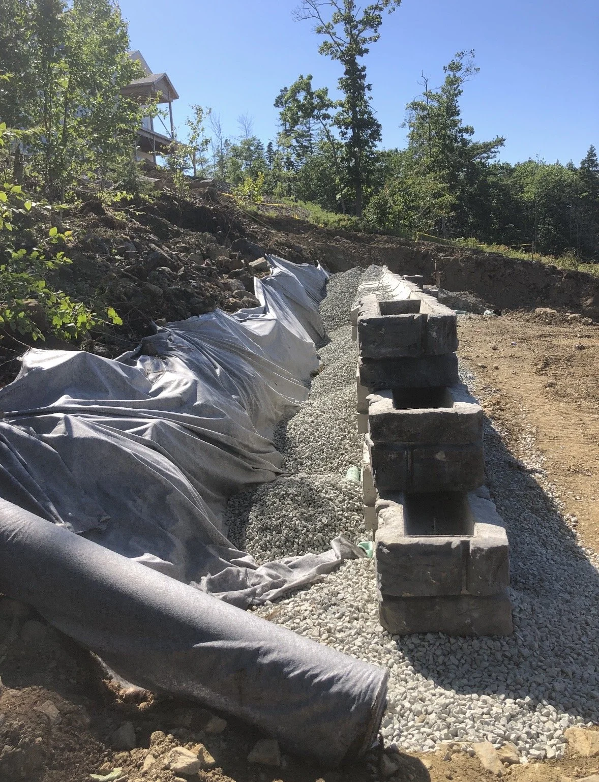 A construction site on a hillside with a row of concrete blocks forming a barrier, covered gravel, and a black plastic sheet on the ground. Forested trees and a house are in the background under a clear blue sky.