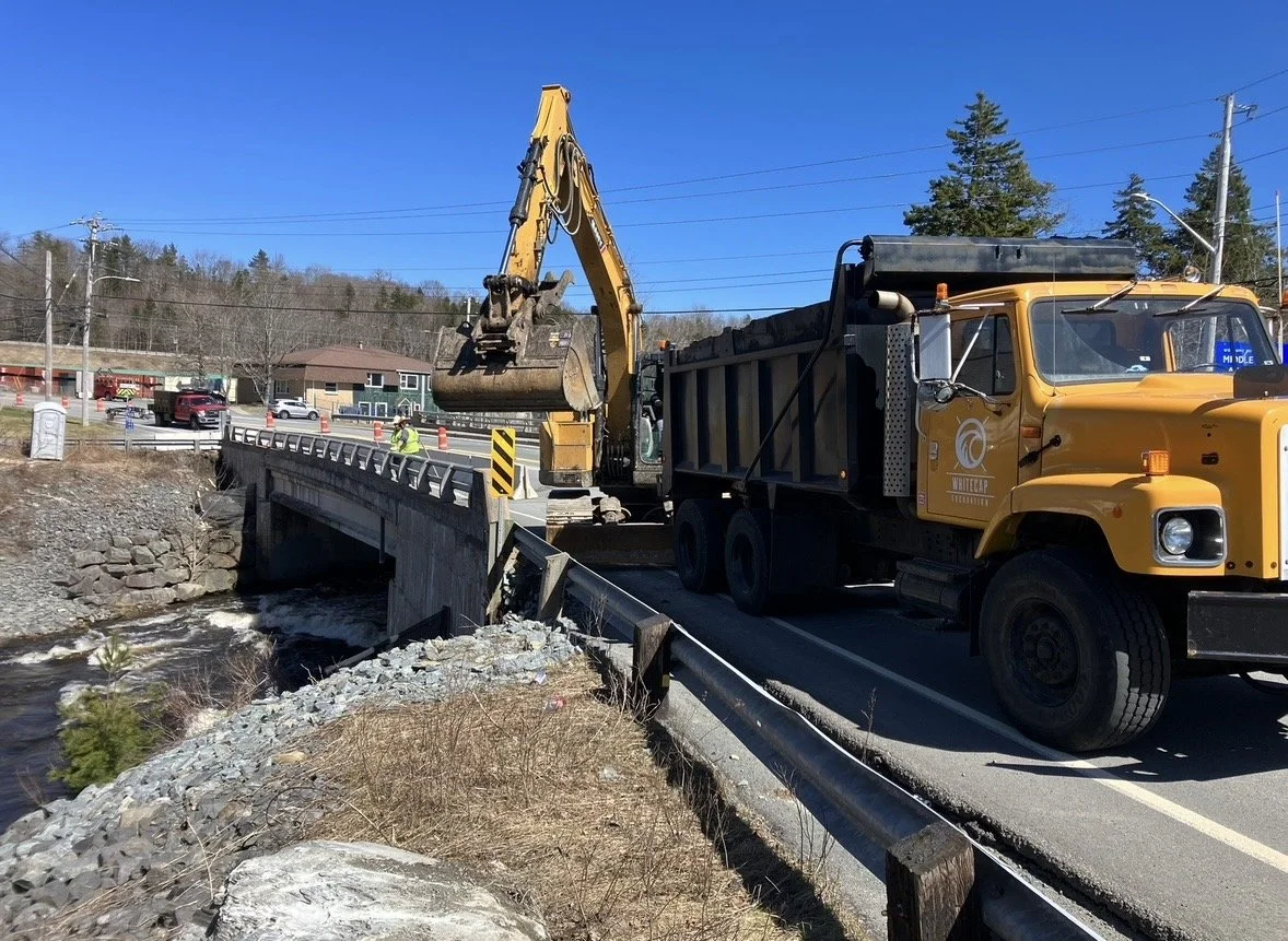 Construction workers working on a bridge repair project with excavator and dump truck on a sunny day.