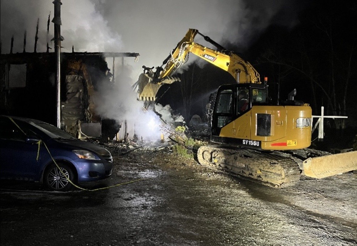 A construction excavator is demolishing a building at night, with smoke and debris around, next to a parked blue car that is cordoned off with yellow tape.