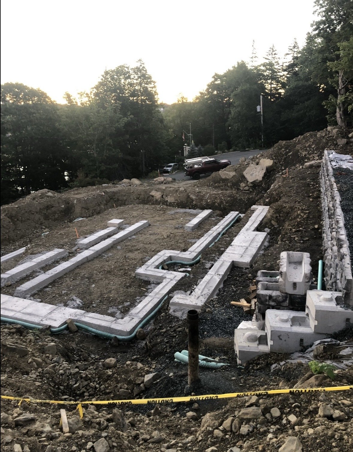 Construction site with concrete blocks and pipes laid out for building foundation, surrounded by dirt and rocks, with caution tape in the foreground, and trees and parked vehicles in the background.