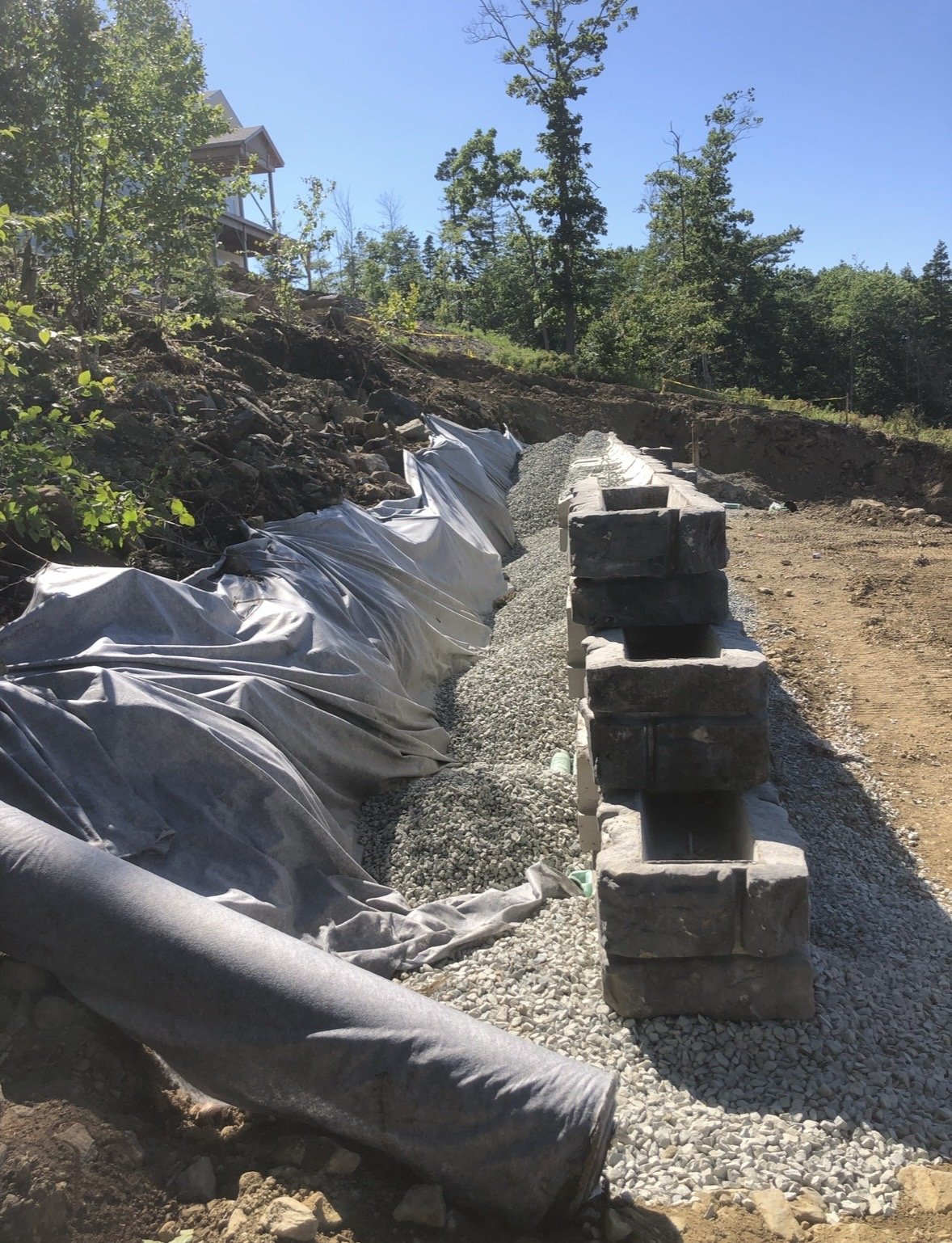 Construction site with a partially built retaining wall made of concrete blocks, gravel, and covered with a tarp on a sloped terrain surrounded by trees and a clear blue sky.