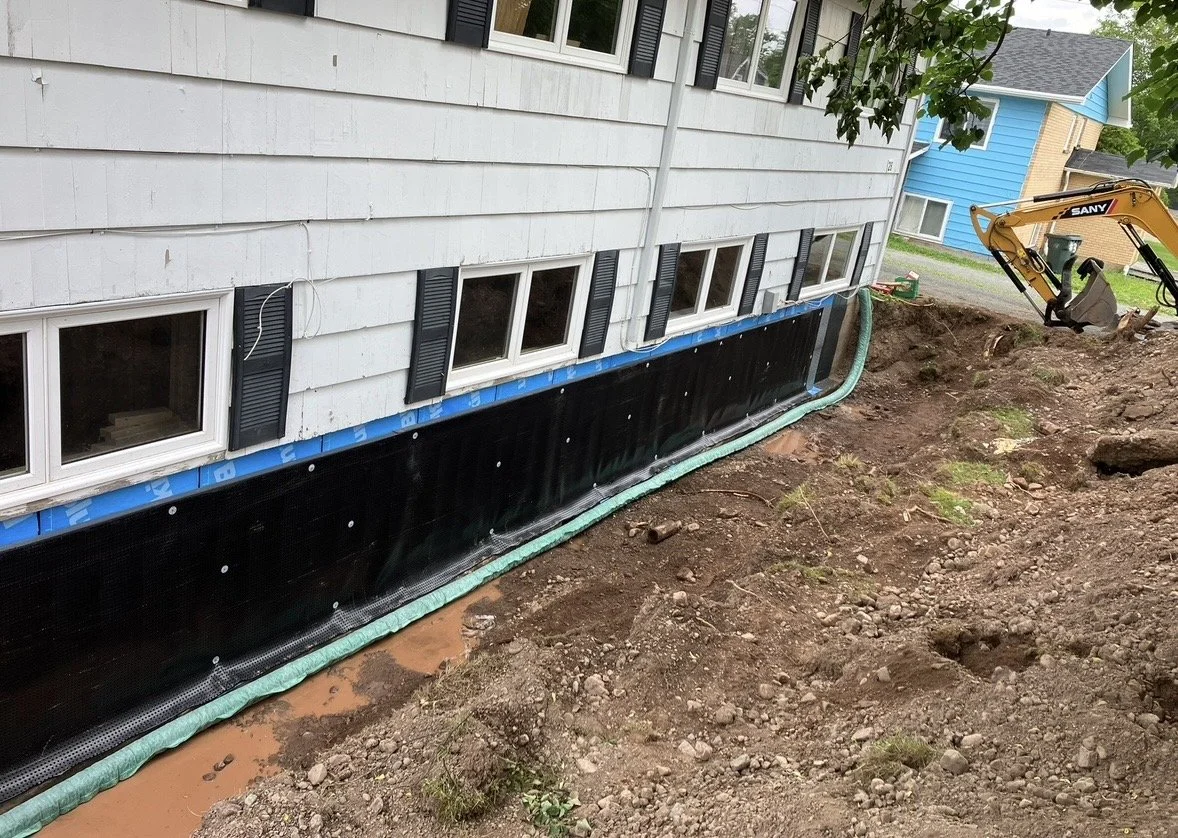 Construction work on the foundation of a house, with black waterproofing membrane installed along the base of the house's exterior wall. The house has white siding and black shutters around the windows. A mini excavator is seen on the right side of the image.