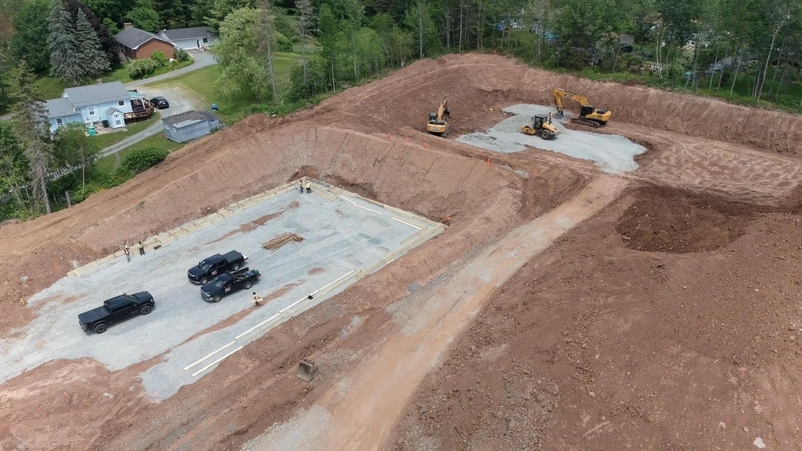 Construction site with excavation equipment and vehicles, preparing foundation for a building, surrounded by residential houses and green trees.
