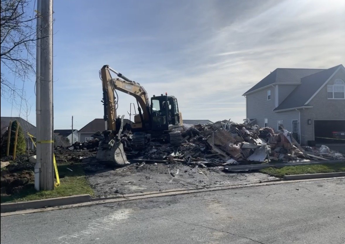 A yellow excavator clearing debris from a demolished house lot in a residential neighborhood.