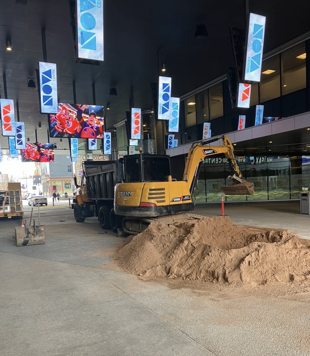 Construction site outside a modern building with multiple digital screens displaying advertisements and a video of hockey players, featuring a yellow excavator digging in a pile of dirt.