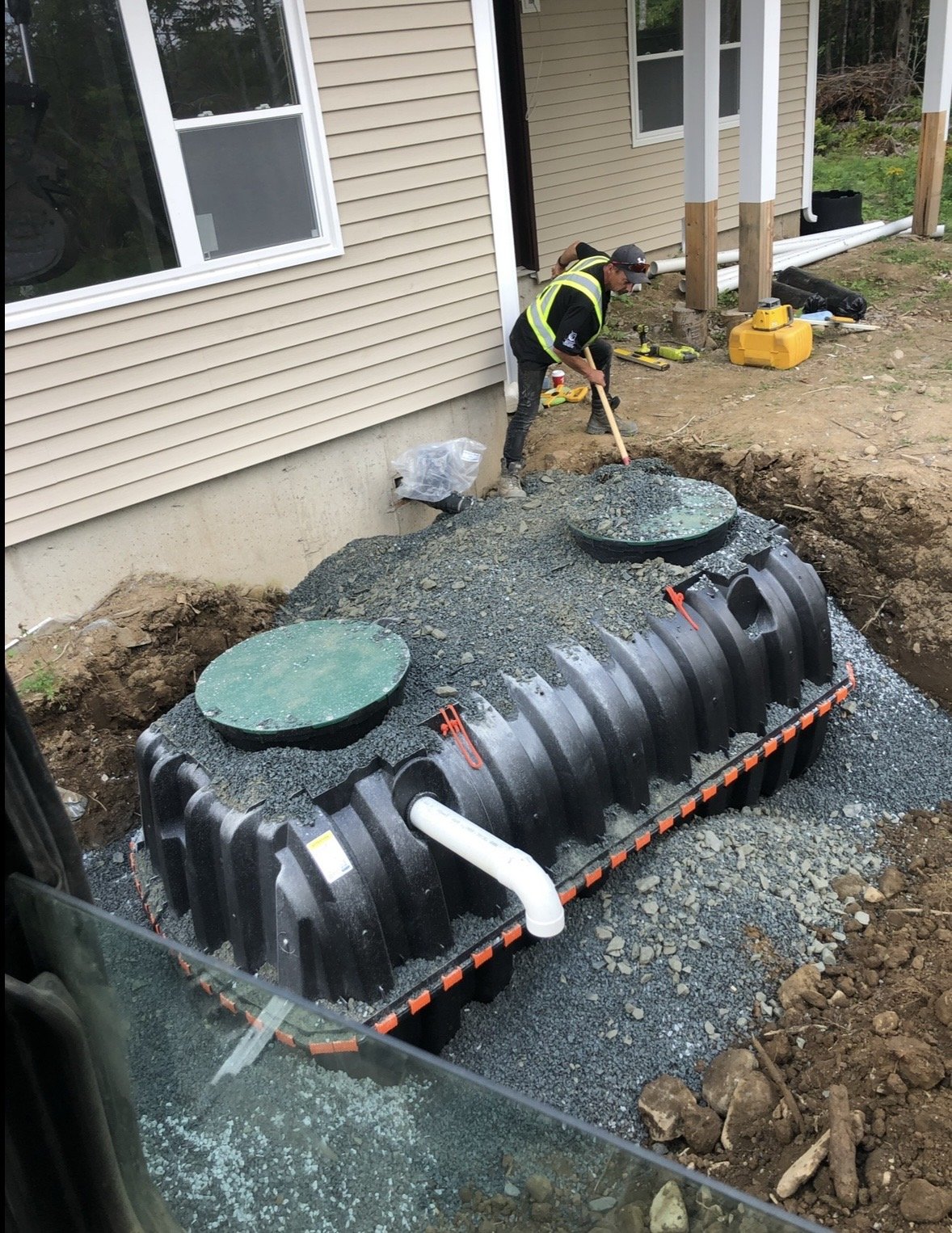 A construction worker in a black jacket with yellow reflective vest digging in the dirt next to a house with beige siding. There is an underground septic tank with two round green access lids and white pipes connected to it.