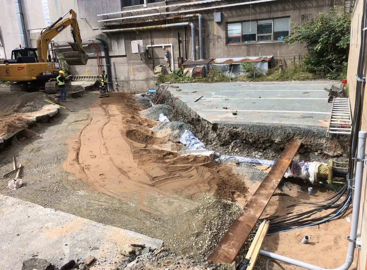 Construction workers and an excavator working on trenching and sidewalk repair at a construction site near a building with parking spaces.