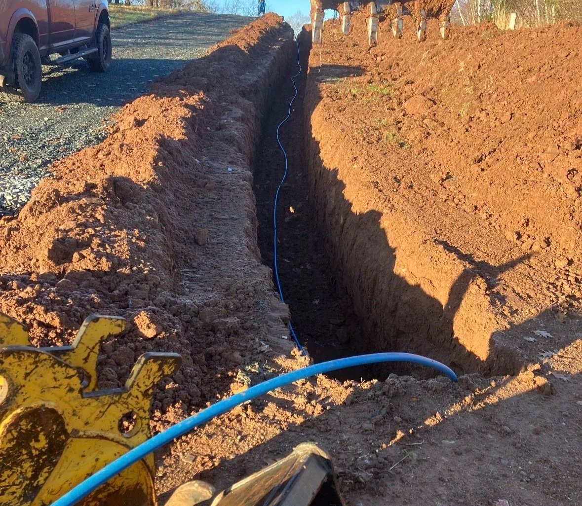 A deep trench in the ground with blue plastic pipes laid inside, part of underground utility installation or repair. A yellow construction tool is partially visible in the foreground. A red vehicle is parked nearby on a gravel surface, with some dirt mounds beside the trench.