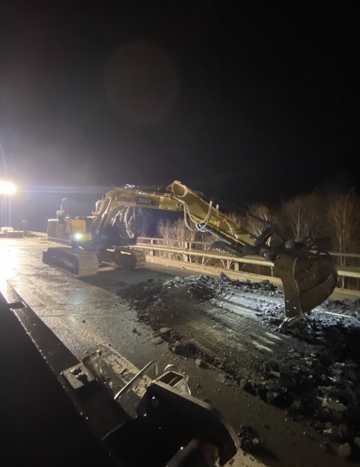 A yellow excavator has crashed into a guardrail on the side of a road at night, with debris and dirt scattered around.