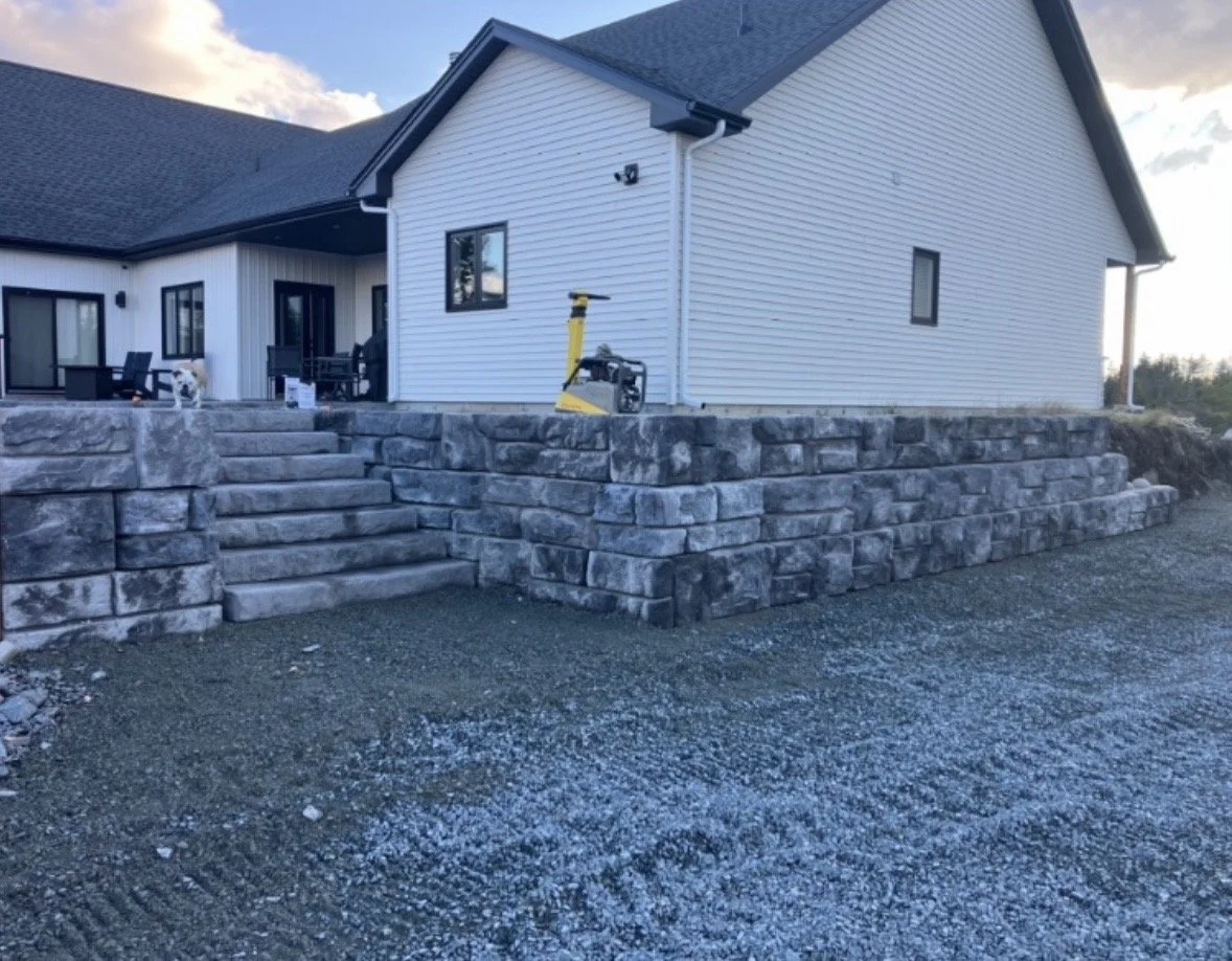 Front of a house with a stone staircase leading up to a porch, white siding, and black window frames. Construction equipment is on the stairs, and the yard is Gravel.