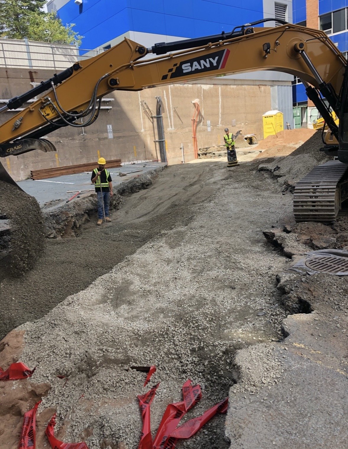 Construction site with excavator and workers wearing safety gear. The ground is being dug up, and there are gravel and dirt piles.