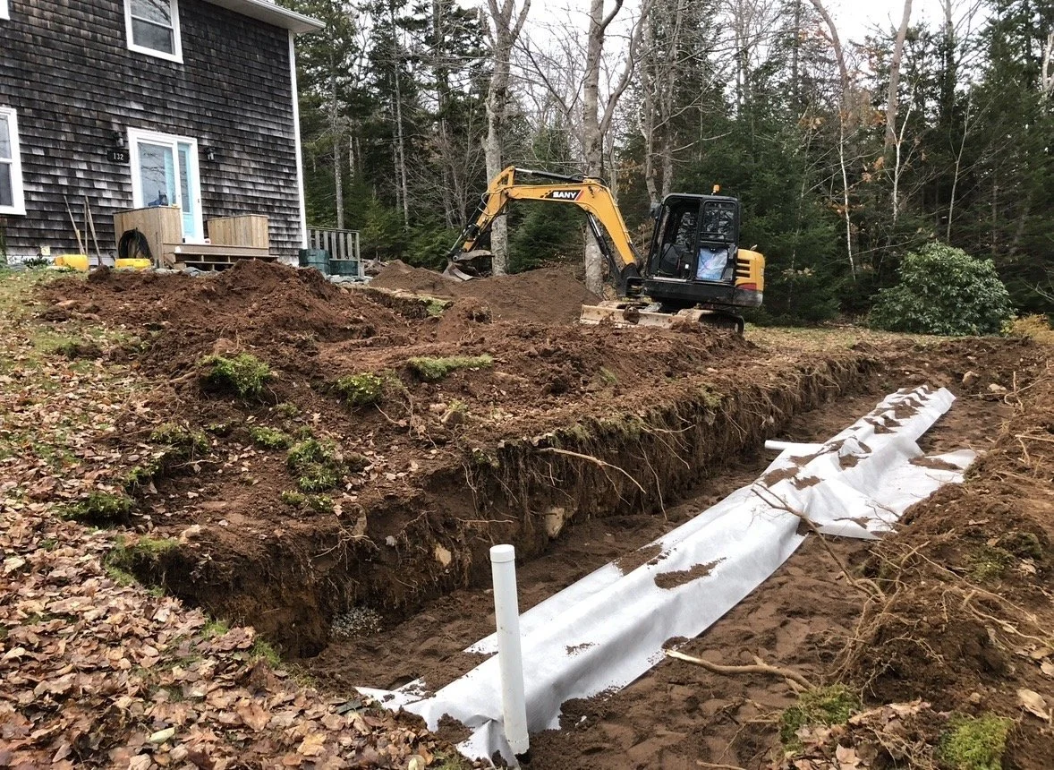A construction site with a small excavator digging near a house. The ground has been excavated, and a white drainage pipe is laid in the trench, covered with a white fabric. There are trees and fallen leaves surrounding the area.