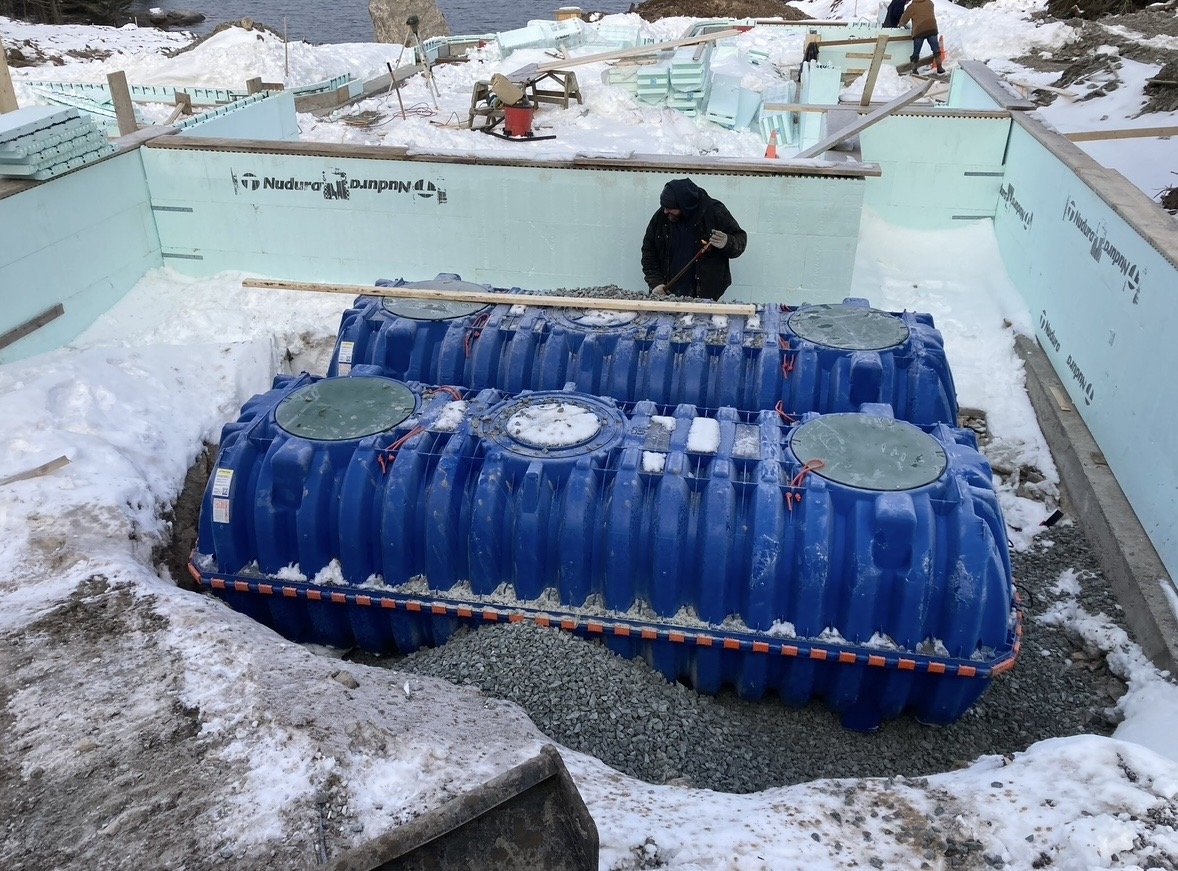 Construction worker standing near a large blue underground septic tank surrounded by snow, with insulated building foundation walls in the background.