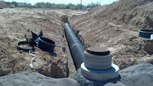 A large drainage pipe in a trench at a construction site with smaller pipes and materials nearby.
