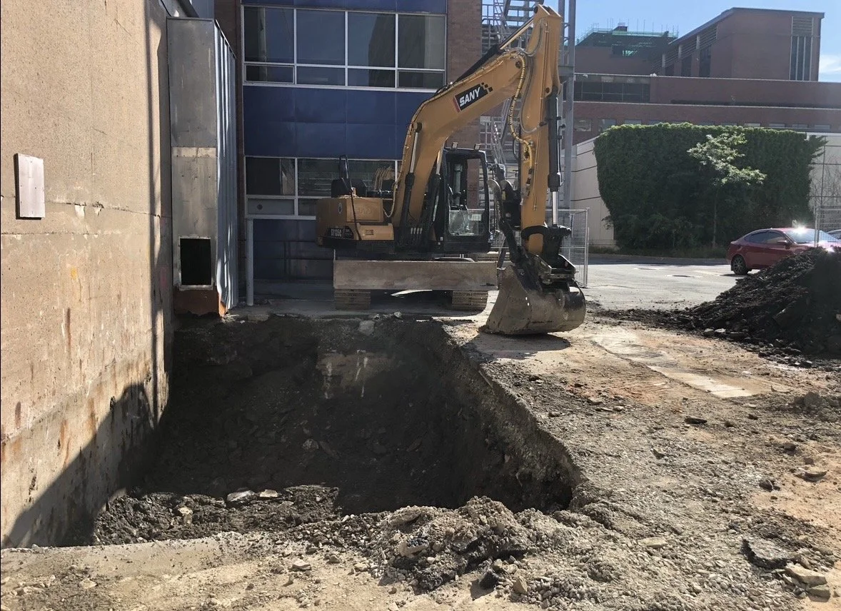 Construction site with a small excavator, a deep hole in the ground, surrounded by dirt and construction materials, in front of a modern building.
