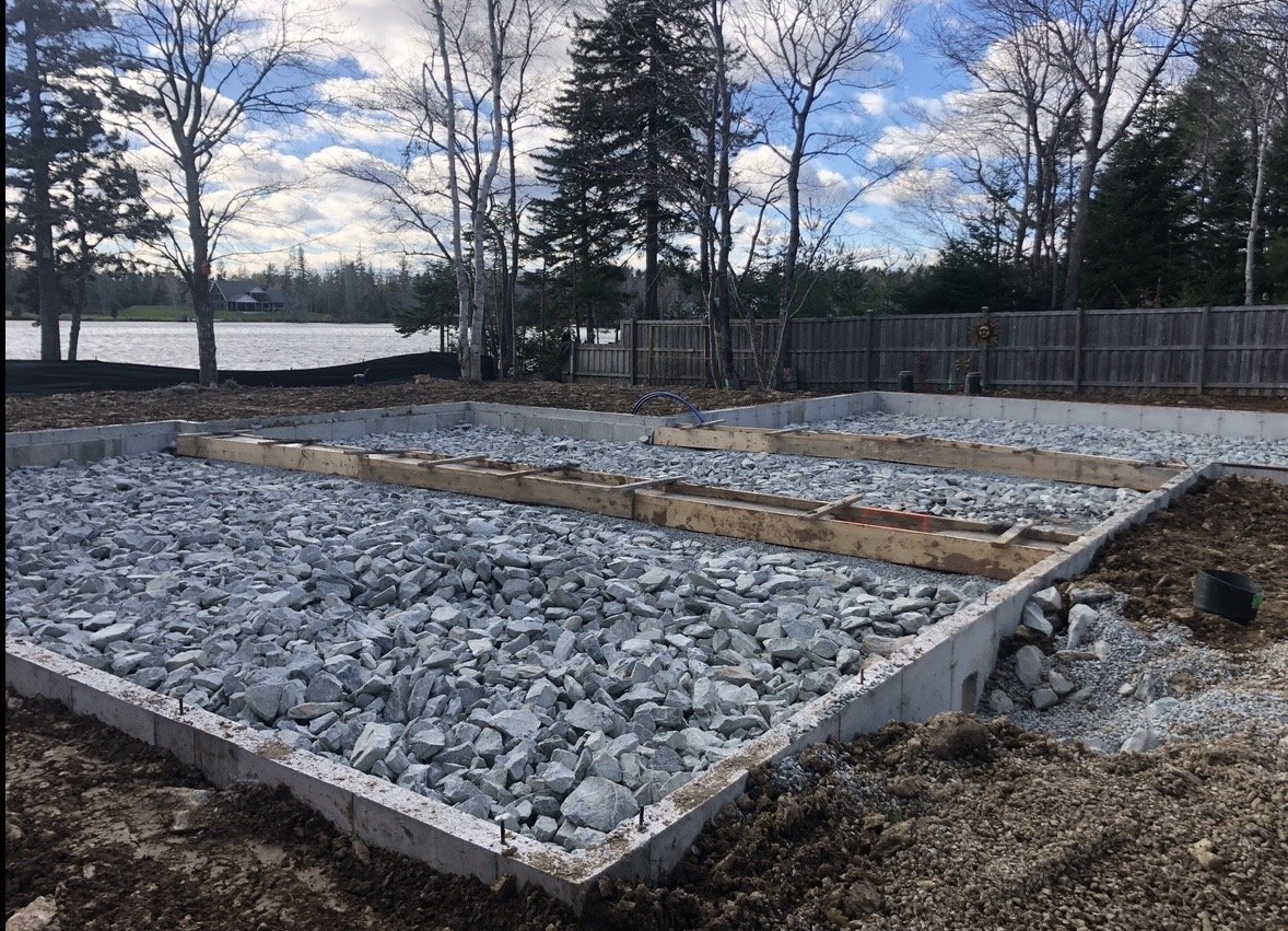 Construction site with concrete foundation and gravel, wooden framing, and a view of trees and a lake in the background.