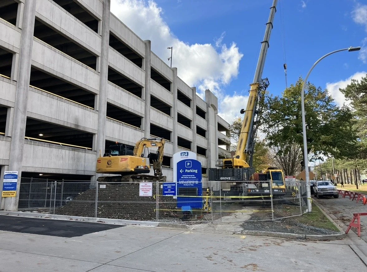 Construction site next to a multi-level parking garage with excavators, a parking sign, safety barriers, and a partly cloudy sky.