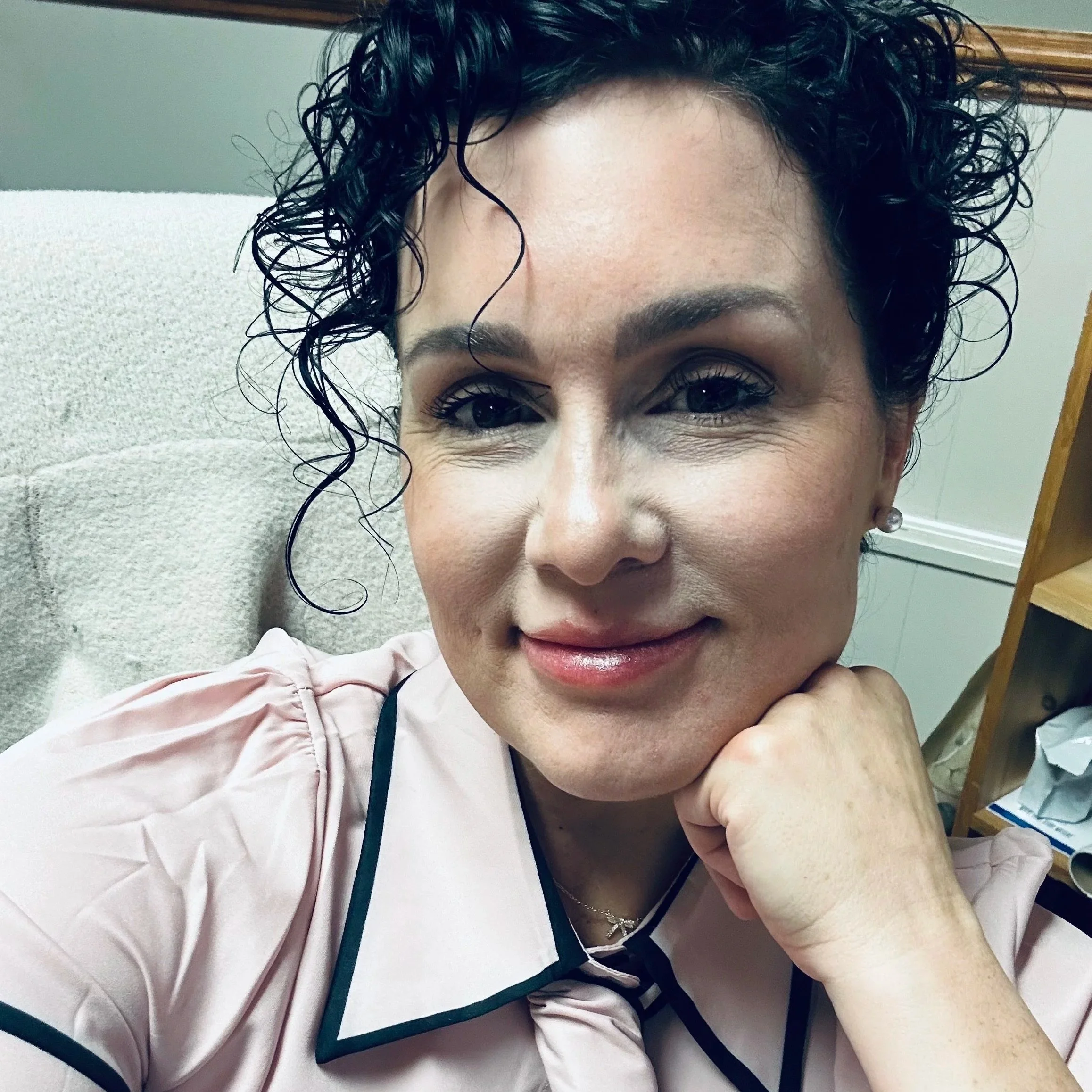 A woman with curly dark hair and wearing a nurse uniform, lying on a hospital bed with her head resting on a pillow, smiling at the camera.