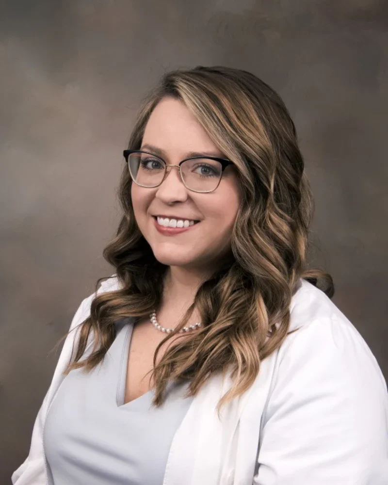 A professional woman with long, curly brown hair wearing glasses, a white lab coat, and a pearl necklace, smiling against a neutral background.