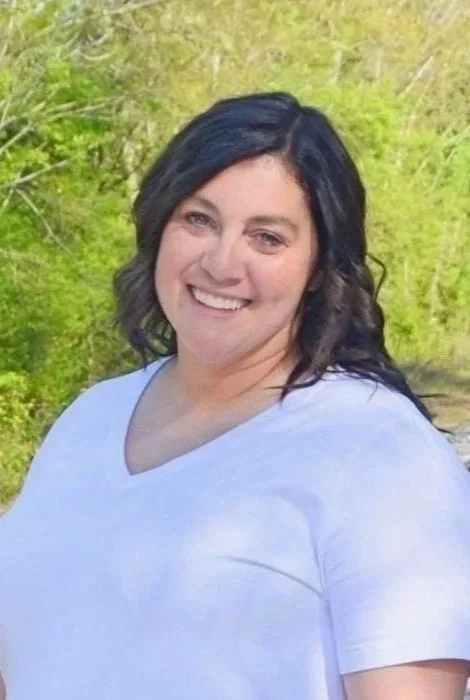 A woman with dark hair smiling outdoors with greenery in the background.