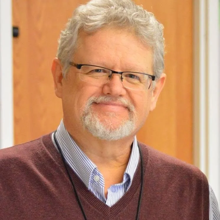 An older man with gray hair, glasses, and a beard, smiling in an indoor setting with a wooden background.