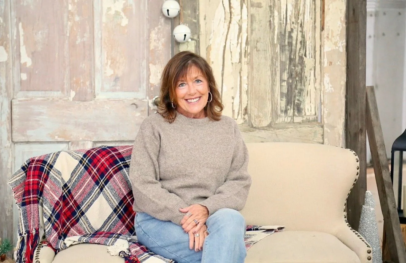 Smiling woman with brown hair sitting on a beige upholstered sofa in a rustic room with weathered wood walls, a plaid blanket, and decorative elements.