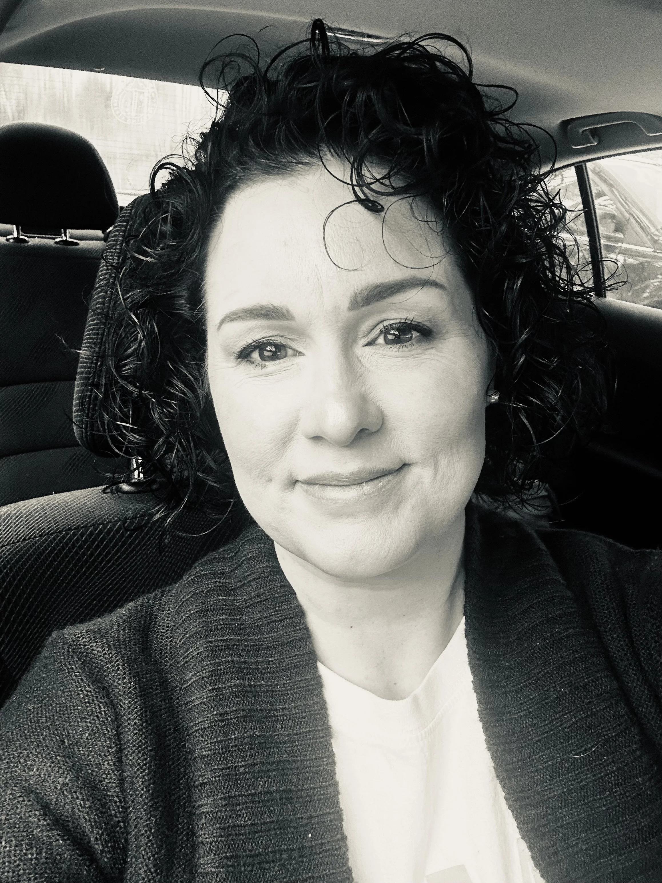 A woman with curly black hair taking a selfie inside a car. She is wearing a light-colored top and a dark cardigan, and is smiling slightly.