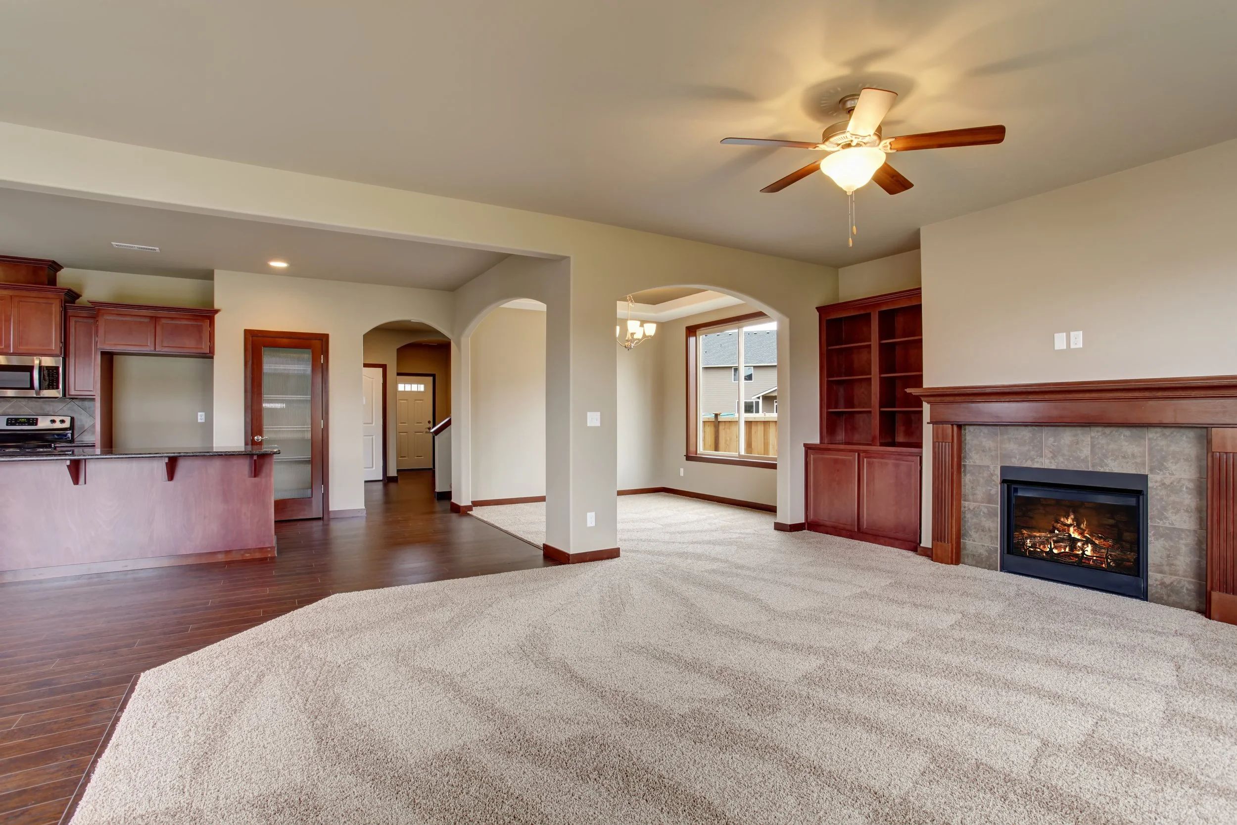 Empty living room with beige carpet, fireplace, wooden built-in shelves, ceiling fan, and large window with view of neighboring houses.