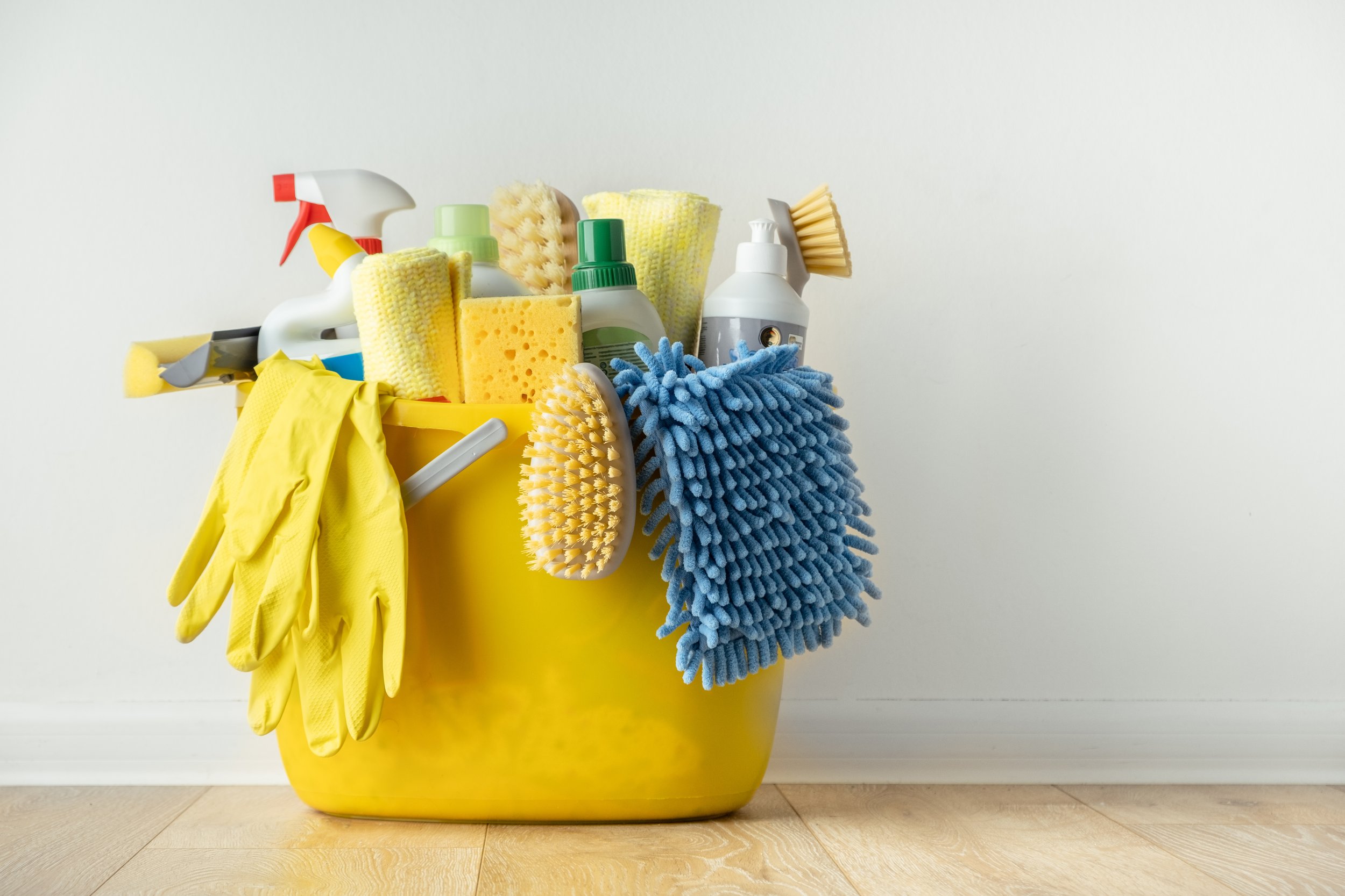 A yellow bucket filled with cleaning supplies including gloves, sponges, brushes, spray bottles, microfiber cloths, and cleaning agents, placed on a wooden floor against a plain white wall.