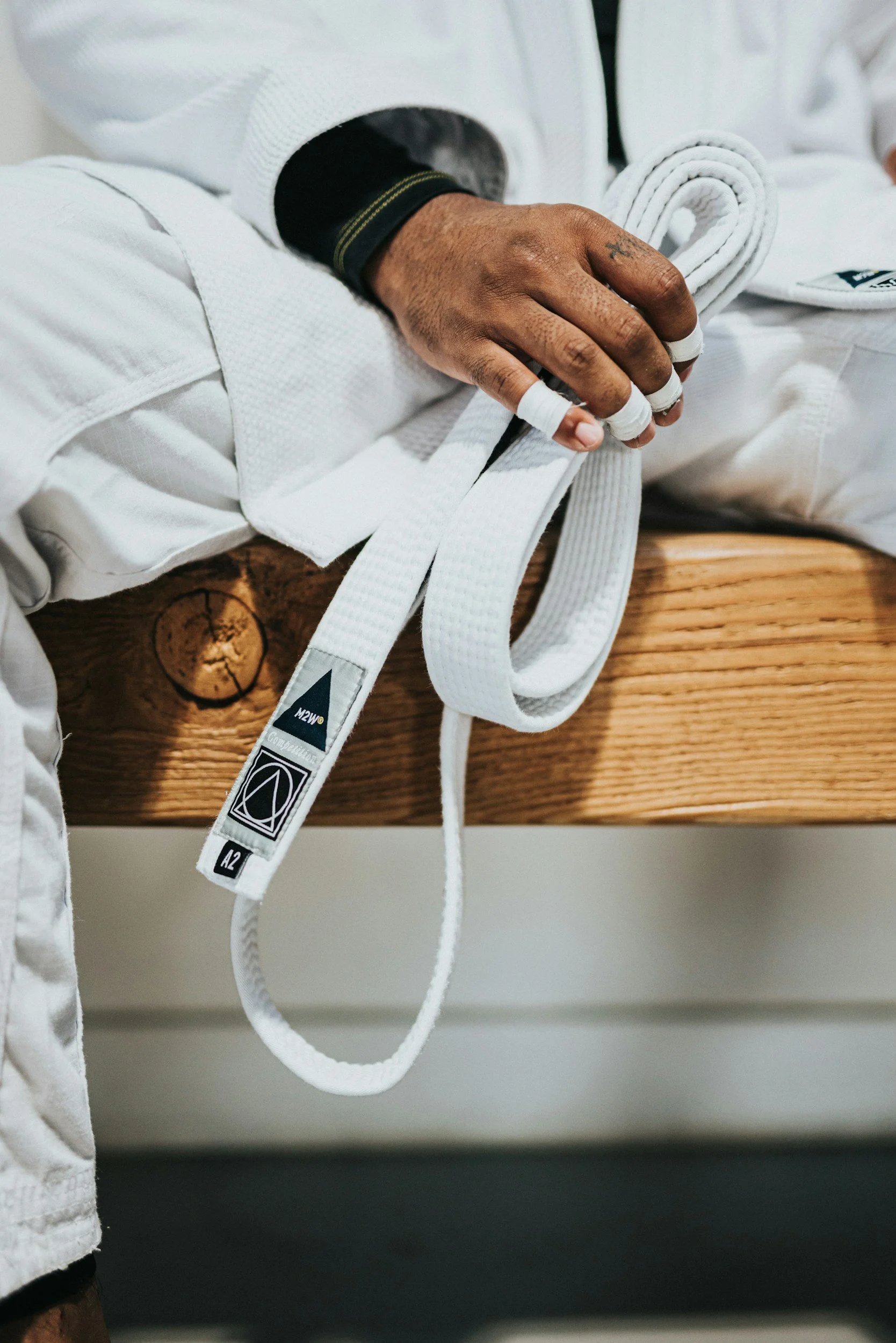 Close-up of a person sitting on a wooden bench with a white martial arts gi and a white belt, holding the belt in their hand with fingers wrapped, showing bandages on fingers.