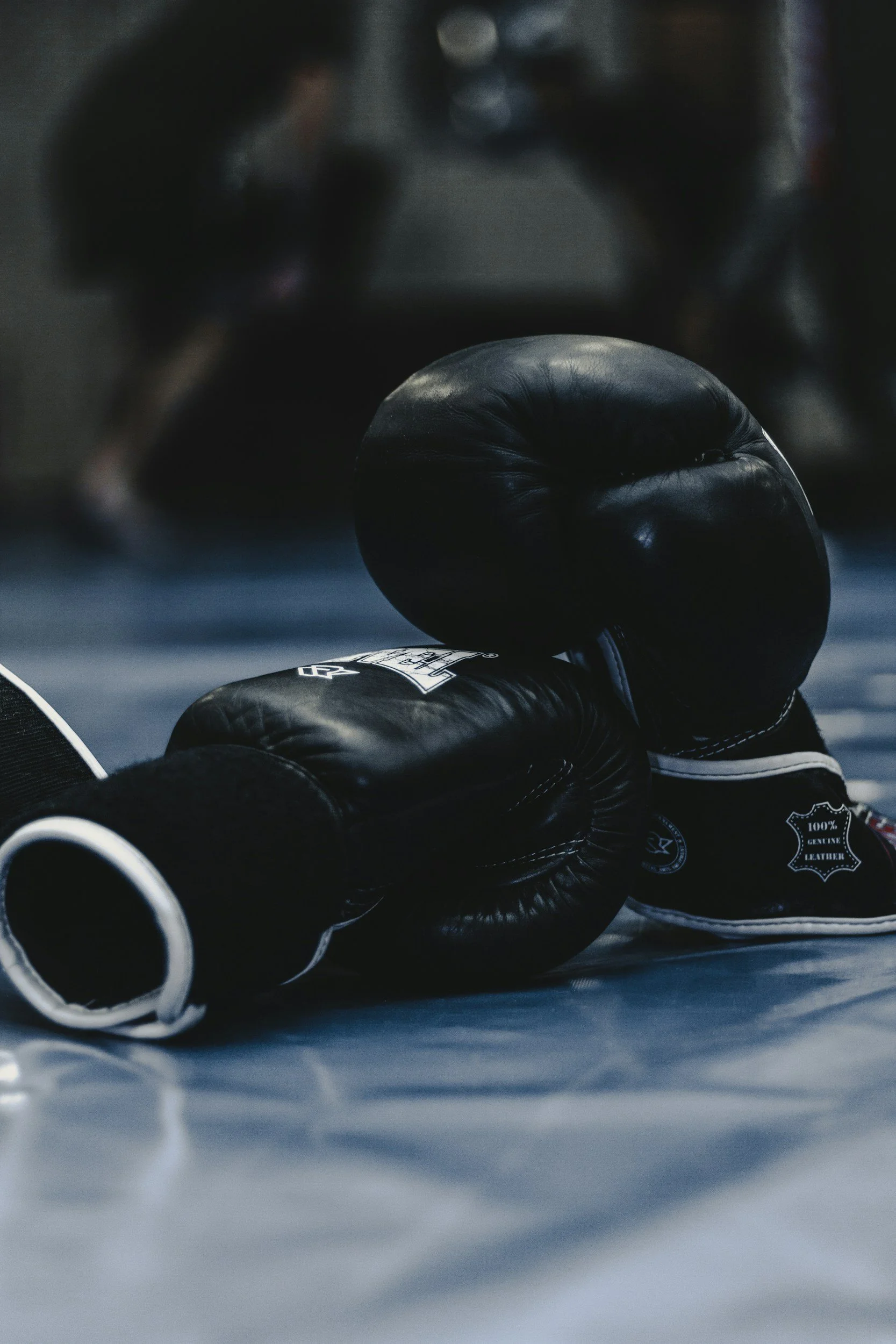 A pair of black boxing gloves on the ground in a boxing ring, with a blurred person in the background.