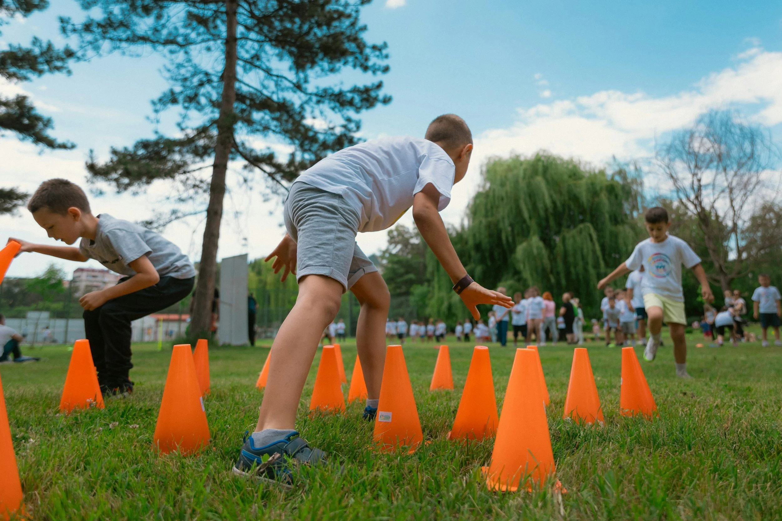 Children participating in an outdoor athletic activity, weaving between orange cones on grass, with trees and a blue sky in the background.