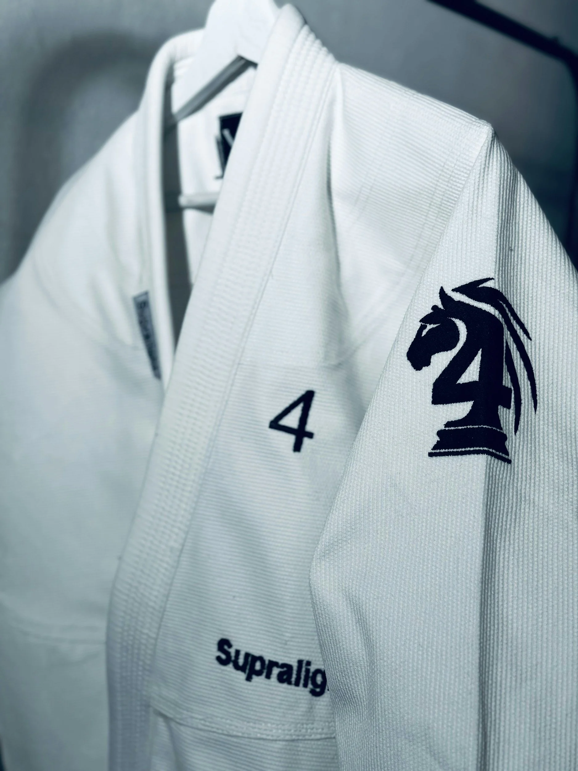 Close-up of a white martial arts gi with a black dragon logo on the sleeve, the number 4 and the word 'Supraling' embroidered on the chest, hanging on a hanger.