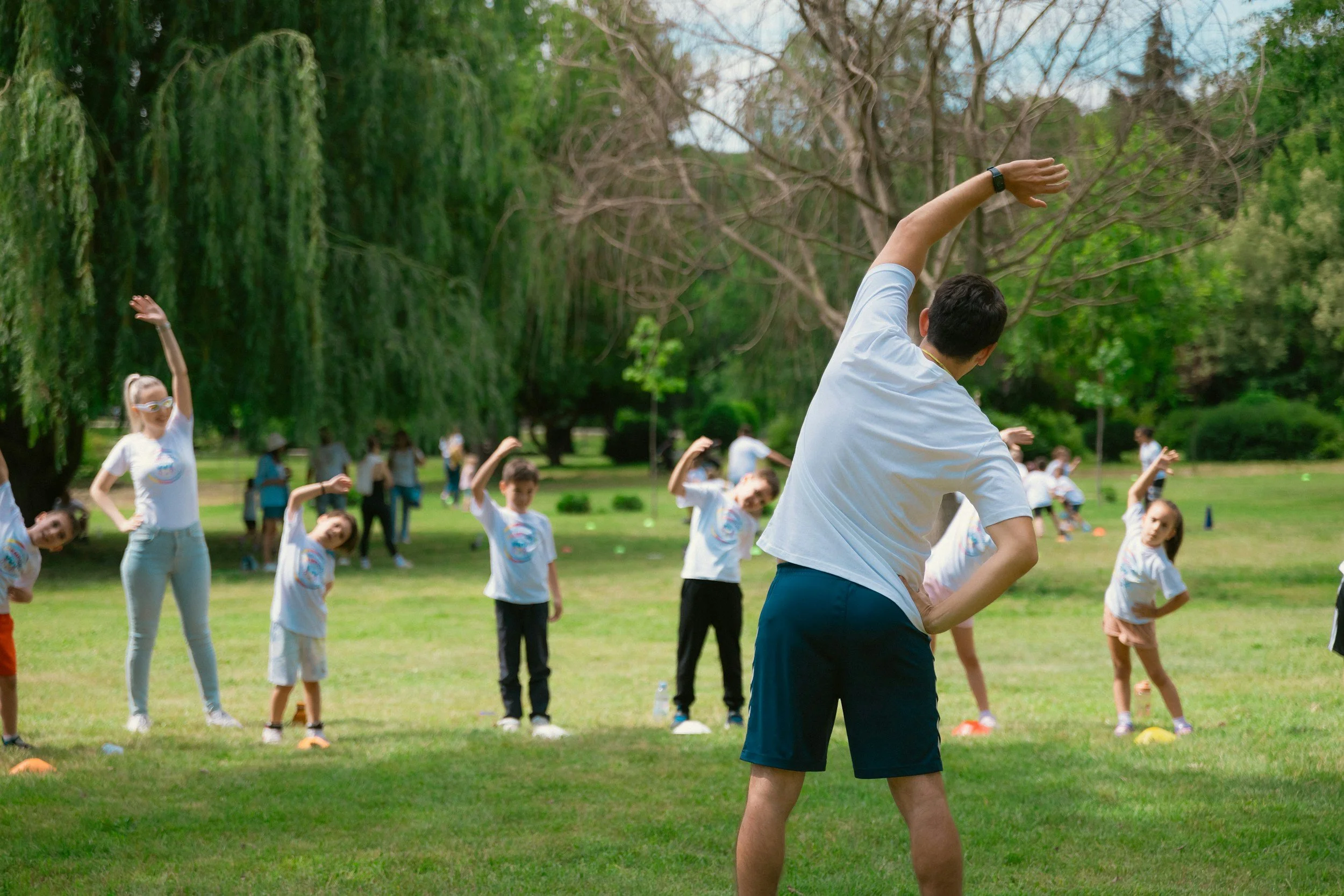 Children participating in an outdoor athletic activity, weaving between orange cones on grass, with trees and a blue sky in the background.