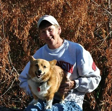 A woman wearing a white baseball cap and a white sweatshirt with red and black text, holding a small tan and white dog, outdoors with tall brown foliage in the background.
