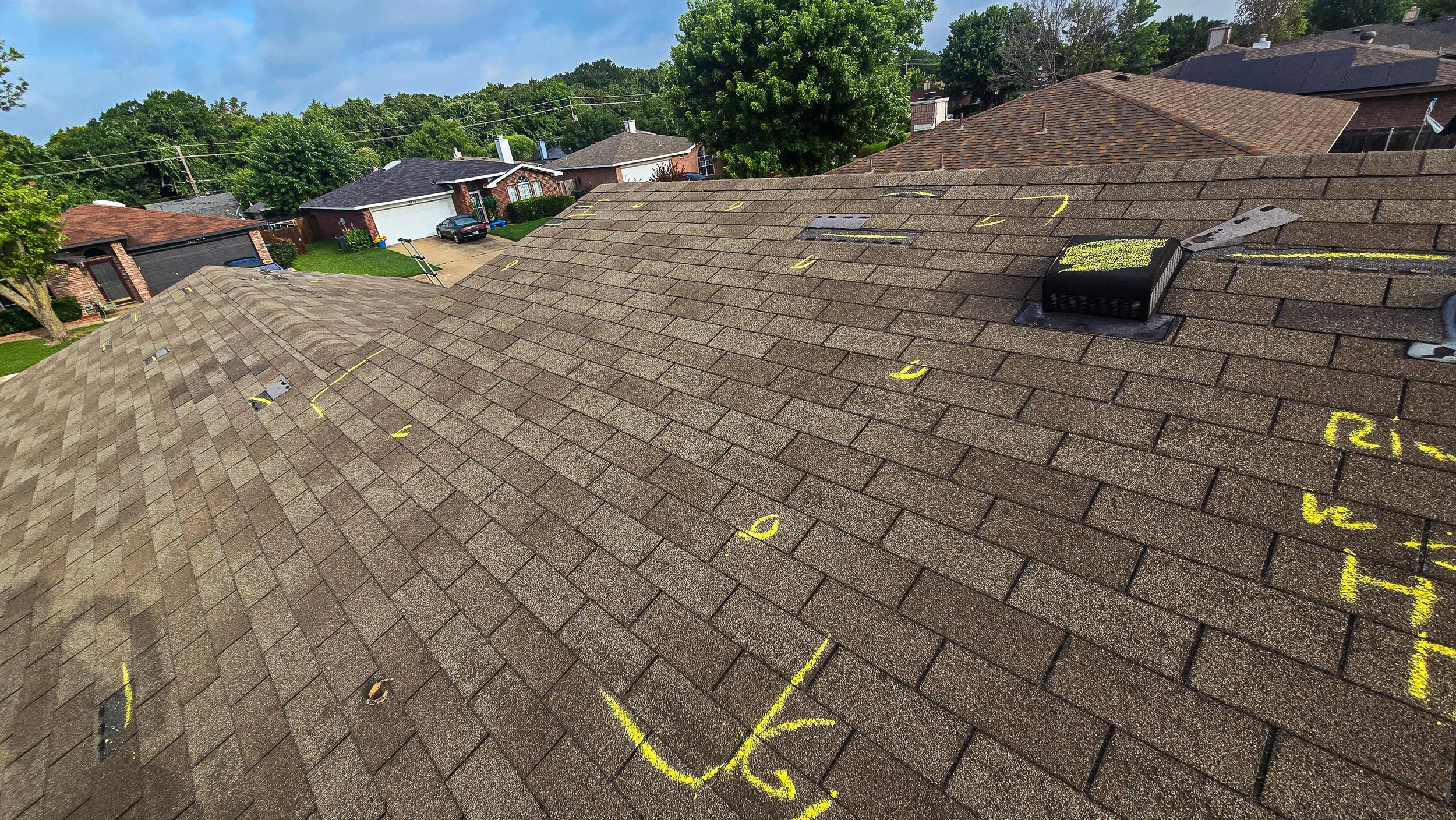 View of a residential roof with brown shingles, marked with yellow spray paint numbers and lines for repair or inspection, and roof vents, with neighboring houses and green trees in the background under a blue sky.