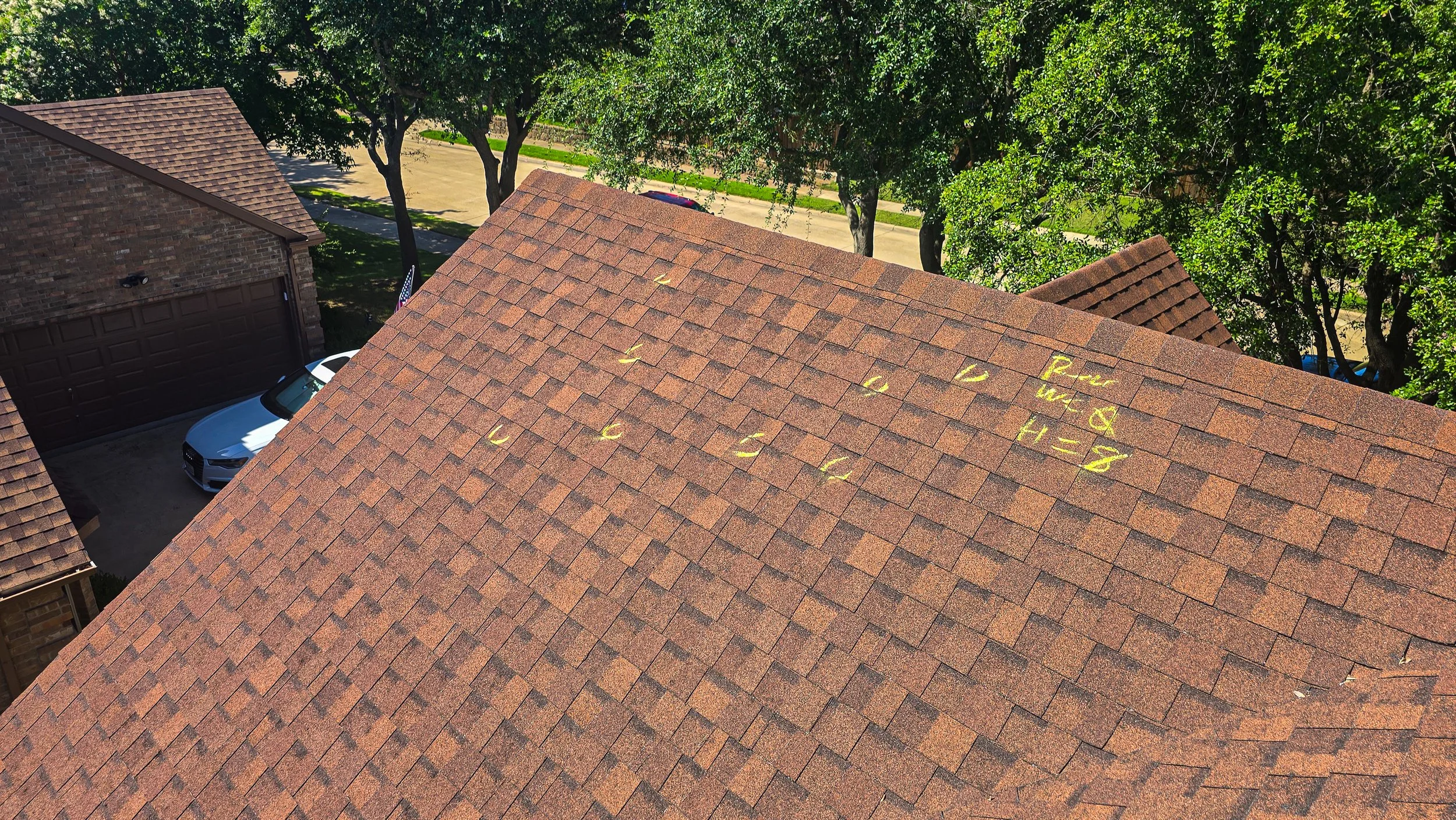 A rooftop with brown shingles, with some numbers and notes written in yellow chalk or paint on it. The background shows trees, a street, and part of a neighboring house.