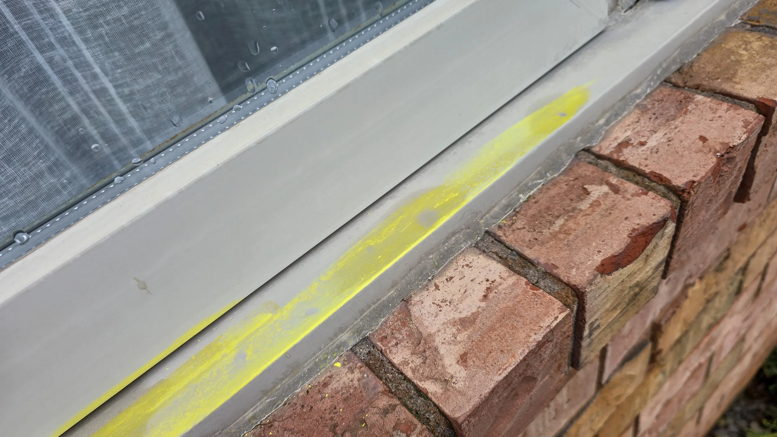 Close-up of a window sill with yellow painter's tape, brick wall, and a window with raindrops on the glass.